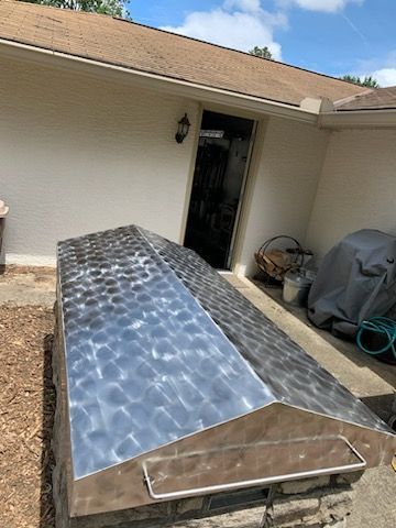 A metal-roofed outdoor stone structure near a house exterior, next to a covered grill and a doorway.