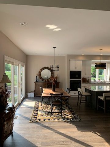 A dining area with a wooden table, chairs on a patterned rug, a round mirror, and a view into a bright kitchen.