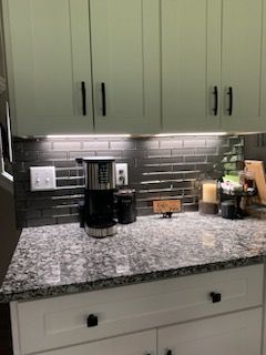 A kitchen coffee station with light cabinets, a speckled granite countertop, and a dark grey tiled backsplash.