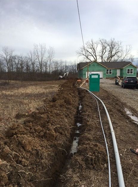 A muddy trench runs through a field next to a house under construction, with pipes, cables, and a portable toilet nearby.