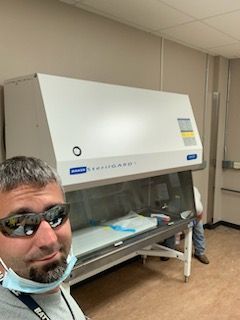 A person wearing sunglasses and a mask takes a selfie in a laboratory next to a white Baker SterilGARD biosafety cabinet.