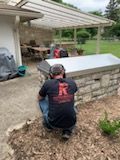 A person in a black t-shirt squats while working on a stone outdoor kitchen counter under a patio cover.