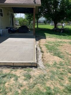 A freshly poured concrete patio with wooden forms, adjacent to a grassy yard and the side of a house.