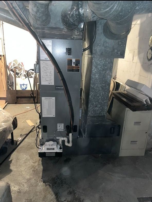 A gray HVAC furnace unit stands in a basement with attached ductwork and a white condensate pump at its base.