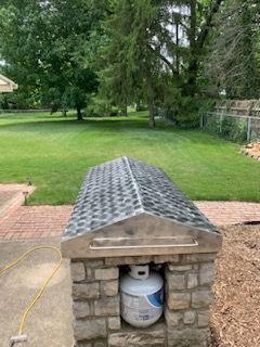 A propane tank sits inside an outdoor stone enclosure with a shingled, gable-style roof in a grassy backyard.