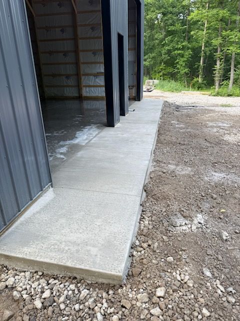 Concrete walkway beside a metal building under construction, with gravel and dirt around it and trees in back.