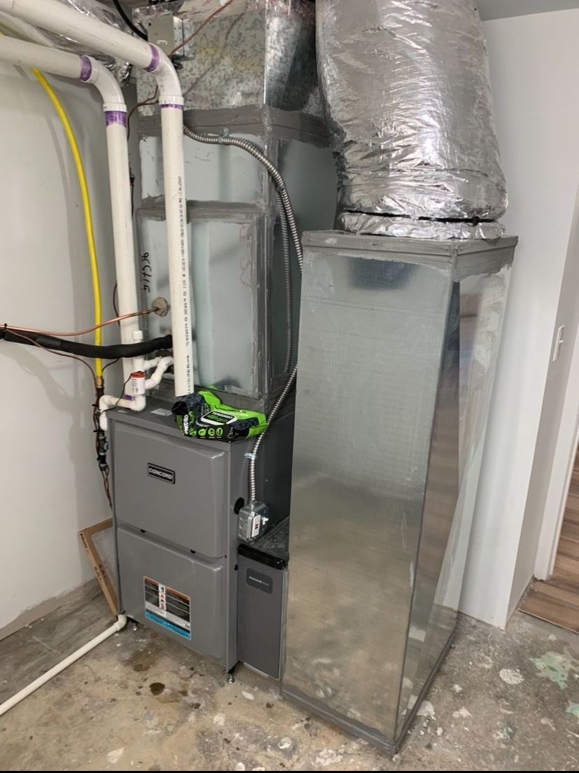 A grey HVAC furnace unit with white PVC intake and exhaust pipes stands in a corner with silver metal ductwork above it.