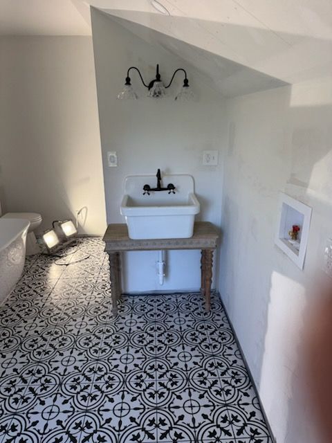 A farmhouse-style sink on a wooden table, featuring a patterned floor, black fixtures, and a wall-mounted light.