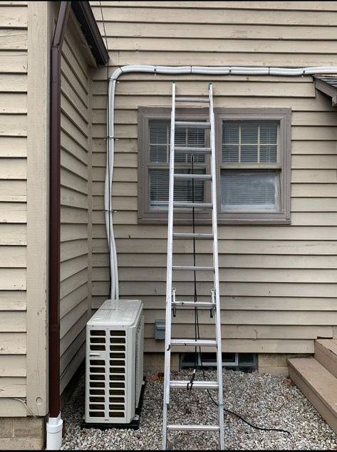 An outdoor HVAC unit sits against a beige house wall next to a tall, extended metal ladder positioned by a window.