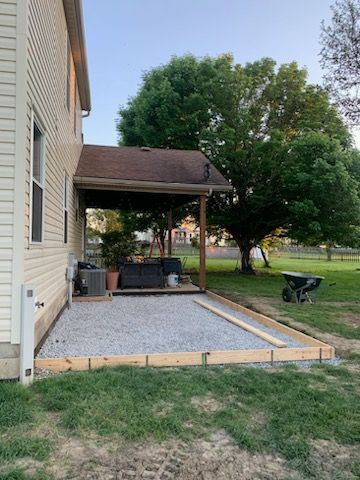 A gravel patio base under construction next to a beige-sided house with a covered porch and a nearby tree.