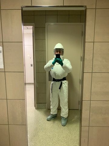A person in a full-body white protective suit, hood, and face shield takes a mirror selfie in a tiled bathroom.