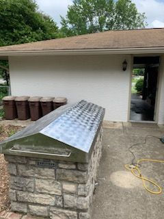 A stone-base grill with a metal roof sits on a patio next to a white house with five brown trash bins in the background.