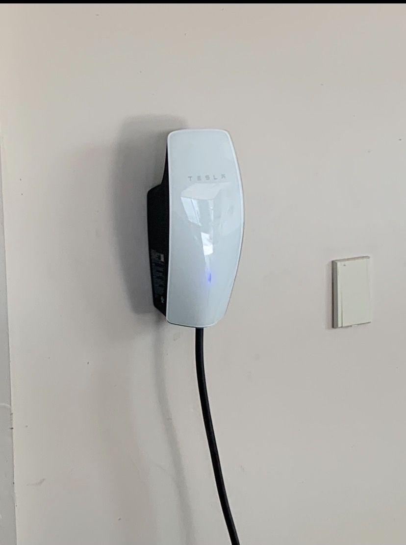 A white Tesla Wall Connector mounted on a plain wall with a black charging cable extending downward.
