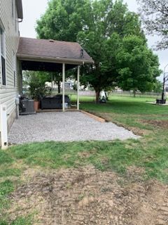 A gravel patio under a covered porch attached to the side of a house, adjacent to a large tree and a grassy yard.