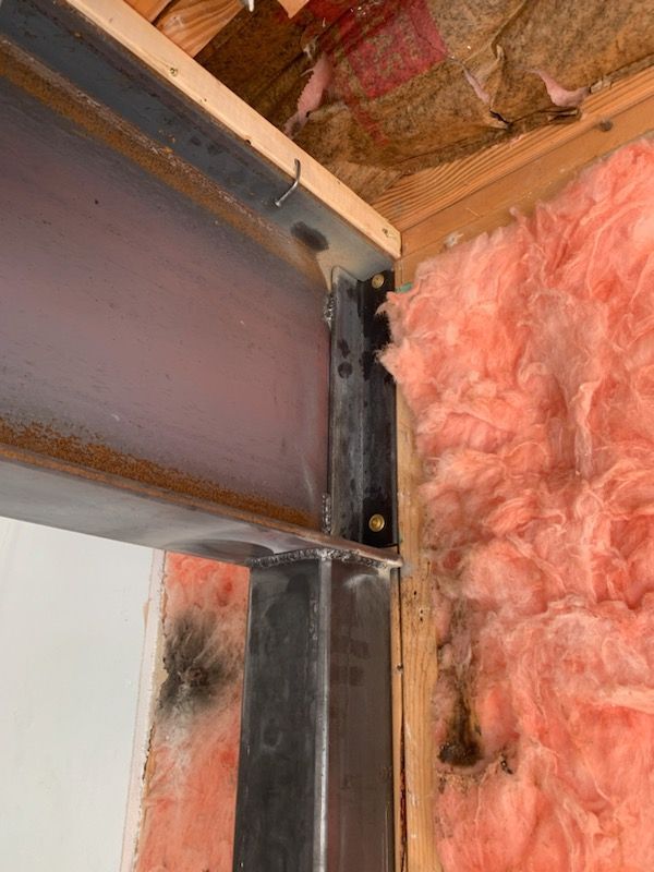 A steel I-beam supported by a vertical steel post against a wall with pink fiberglass insulation.