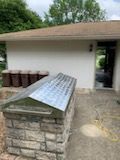 A stone structure with a metal peaked roof sits on a patio next to a house exterior with a row of potted plants.