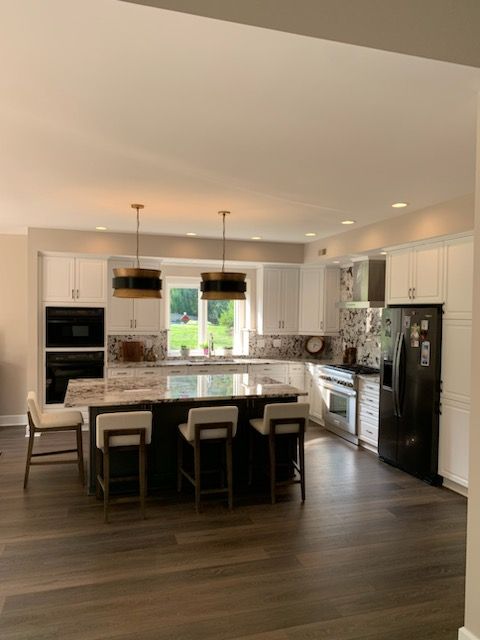 A bright, modern kitchen featuring white cabinets, a dark island with three stools, pendant lighting, and stainless steel.