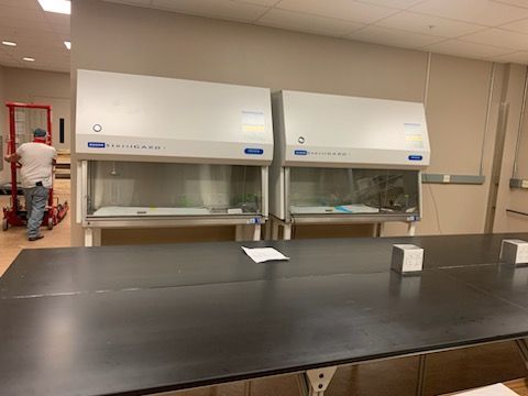 Two biological safety cabinets sit on a lab bench near a person operating a red equipment lift in a bright room.