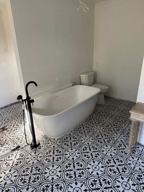 A white freestanding bathtub with a black faucet sits on a black-and-white patterned tile floor next to a toilet.