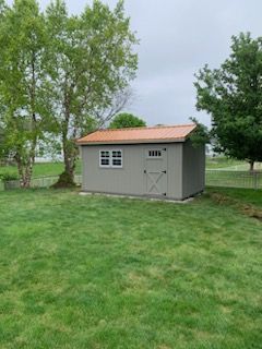 A gray storage shed with a copper-colored metal roof and a single door and window, situated in a grassy backyard.