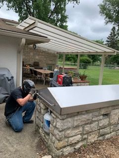 A welder in protective gear works on a stainless steel grill cover on a stone island under a patio cover.