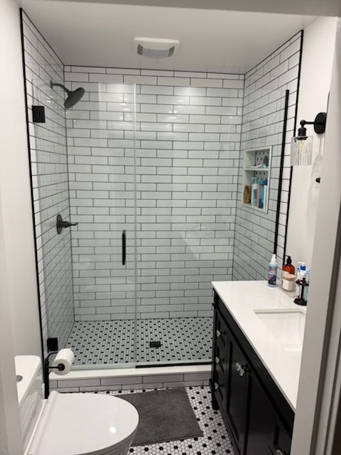 A modern bathroom with a glass shower enclosure, white subway tile walls, patterned black and white floor, and vanity.