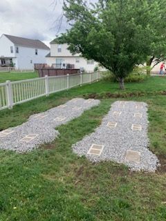 Two strips of gravel with rectangular concrete blocks set into a green lawn near a white fence and a large tree.
