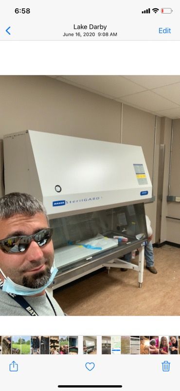 A man wearing a mask and sunglasses takes a selfie in a room with a large, white laminar flow hood in the background.