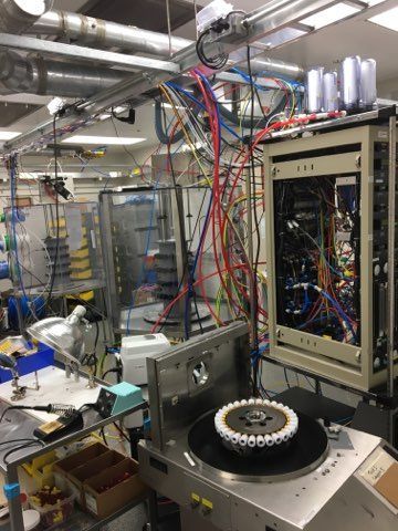A laboratory workspace featuring a round sample carousel, complex wiring, and server equipment in a cluttered room.