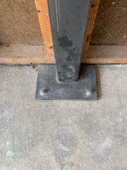 A gray metal support post with a rectangular base plate bolted to a concrete floor in front of wooden wall framing.