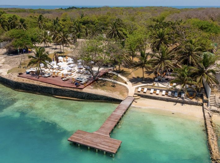 Playa de arena con agua turquesa, palmeras y edificios de piedra.