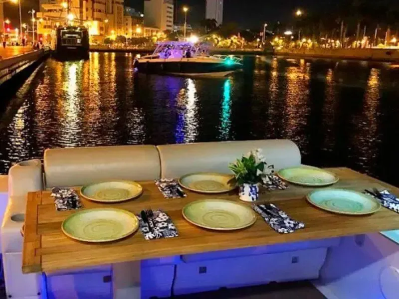 Mesa de barco preparada para una cena nocturna, entorno frente al mar con luces de la ciudad y reflejos en el agua.