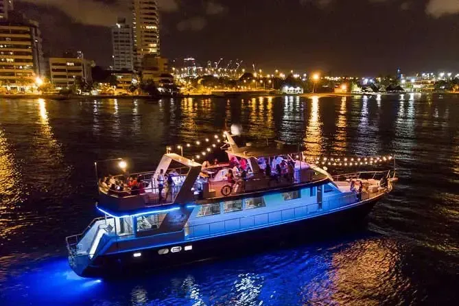 Vista nocturna de una fiesta en barco sobre el agua, con las luces de la ciudad al fondo.