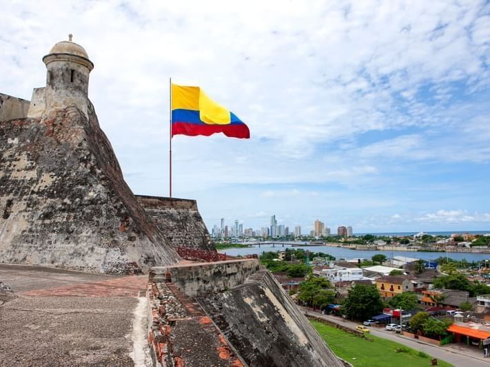 La bandera colombiana ondea sobre la fortaleza; al fondo se ve el horizonte de la ciudad y el agua.