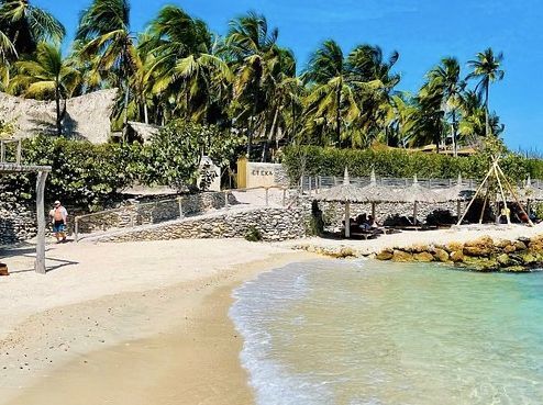 Playa de arena con agua turquesa, palmeras y edificios de piedra.