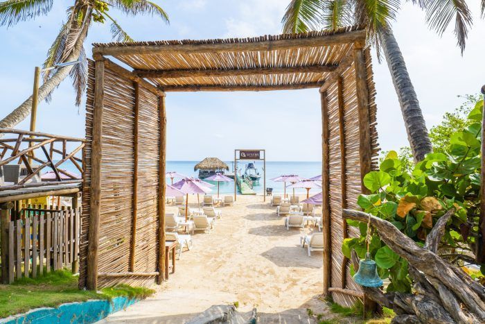 Un arco de paja enmarca una escena playera: arena blanca, tumbonas con sombrillas y agua turquesa.