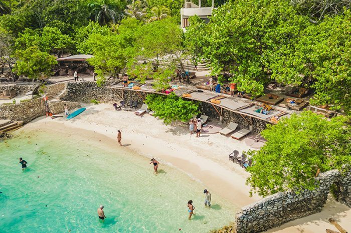 Complejo turístico frente al mar con edificios con techo de paja, exuberante vegetación y tranquilas aguas turquesas.