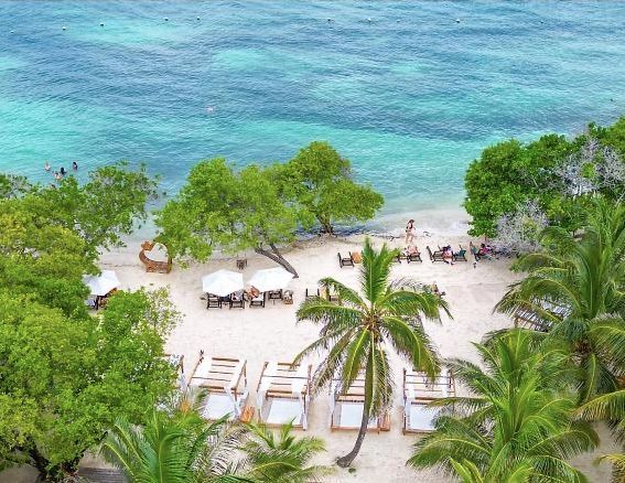 Playa con cabañas, palmeras y aguas turquesas. Orilla de arena con tumbonas bajo estructuras cubiertas.