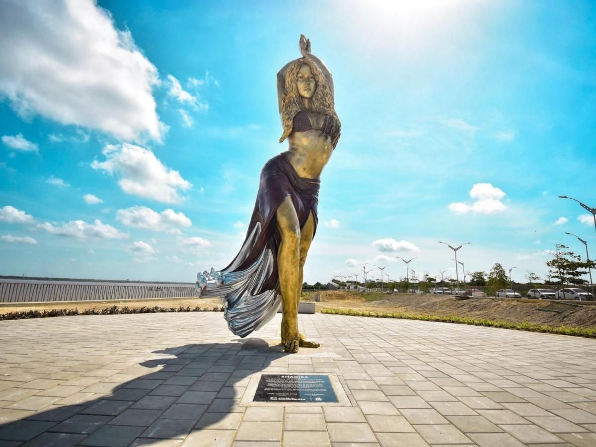 Estatua de una mujer bailando, con los brazos en alto. Escenario al aire libre, cielo azul y plaza pavimentada.