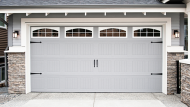 A light gray, single-car garage door with arched, paneled windows and black hardware on a house with stone-accented walls.