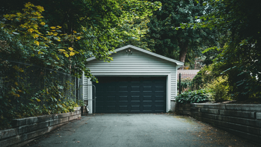 A white garage at the end of a tree-lined driveway with a dark double door.