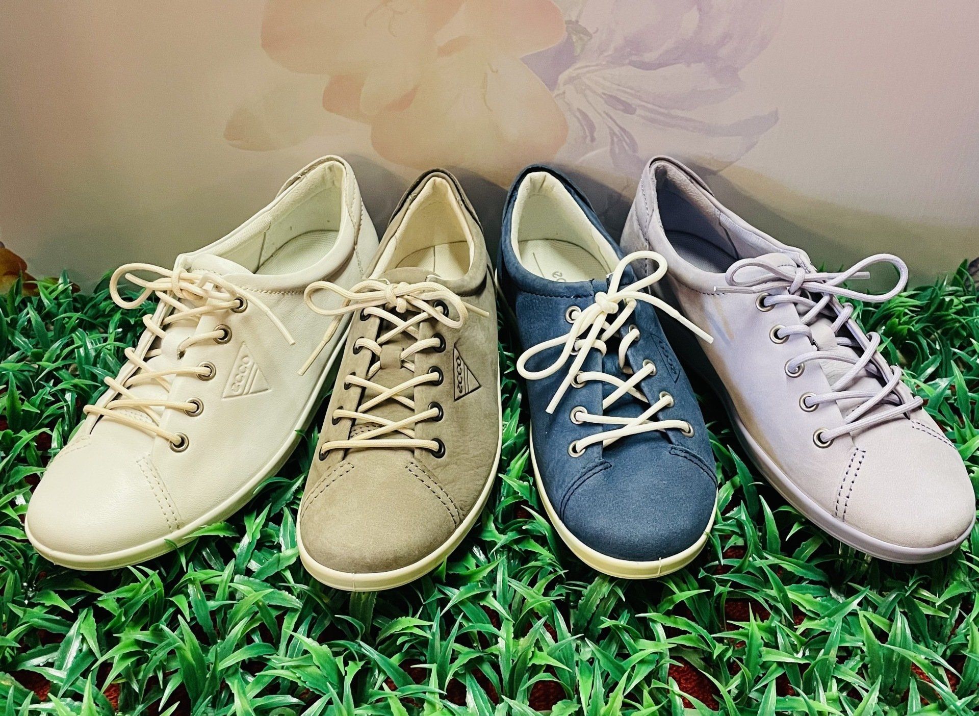 Quattro paia di scarpe da ginnastica con i lacci, di colori diversi: crema, grigio-marrone, blu denim e lavanda, su erba verde finta.