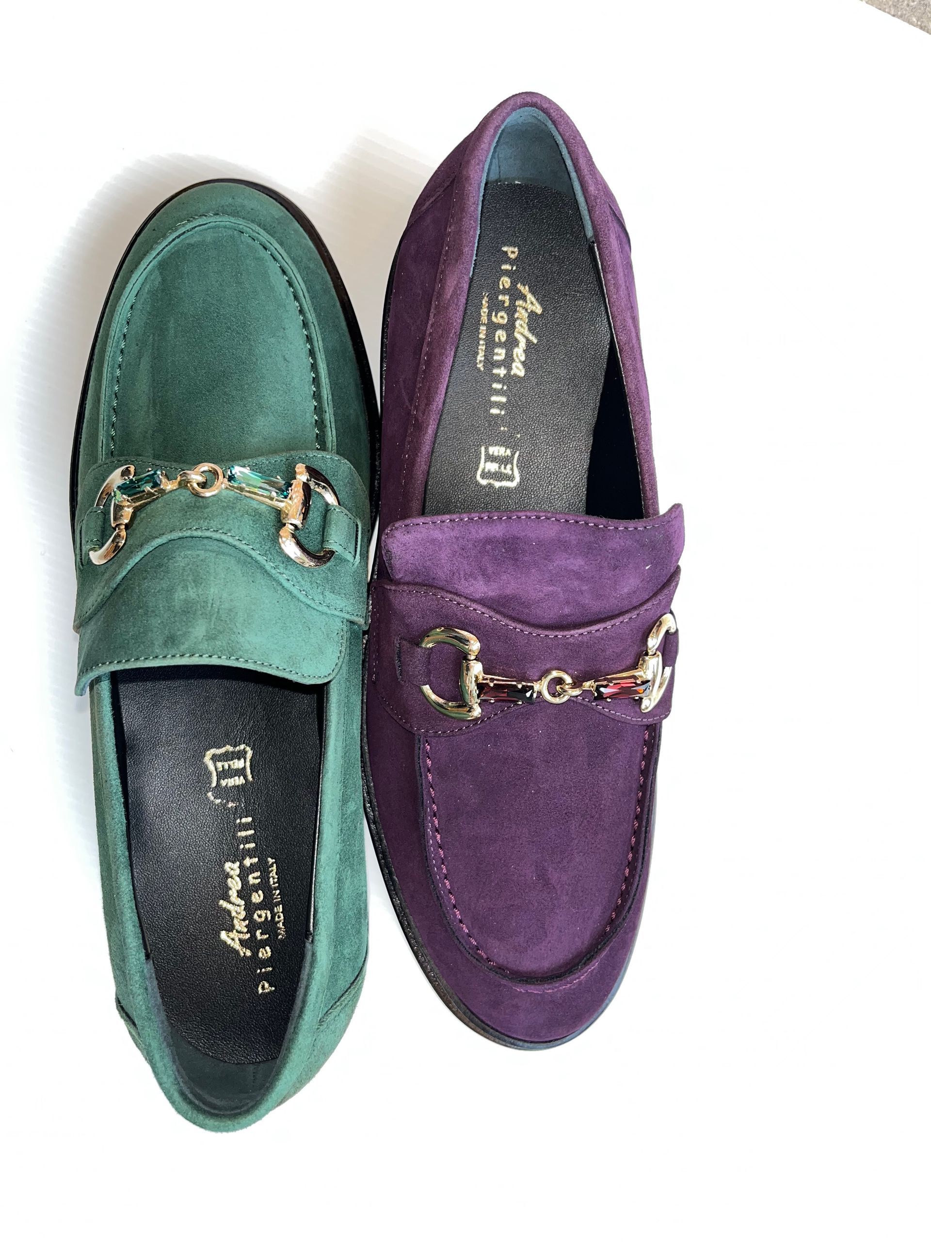 Un paio di mocassini in pelle scamosciata, uno verde e uno viola, con dettagli color oro su superficie bianca.