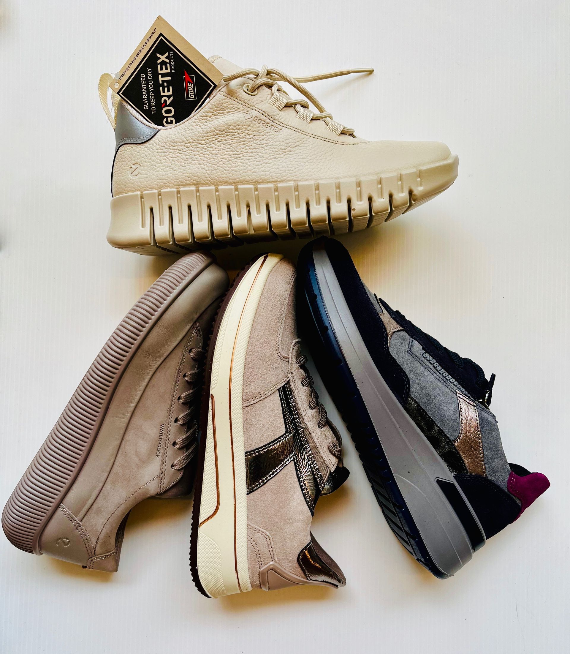 Quattro paia di scarpe da ginnastica beige e grigie disposte su una superficie bianca. Una ha l'etichetta in Gore-Tex.