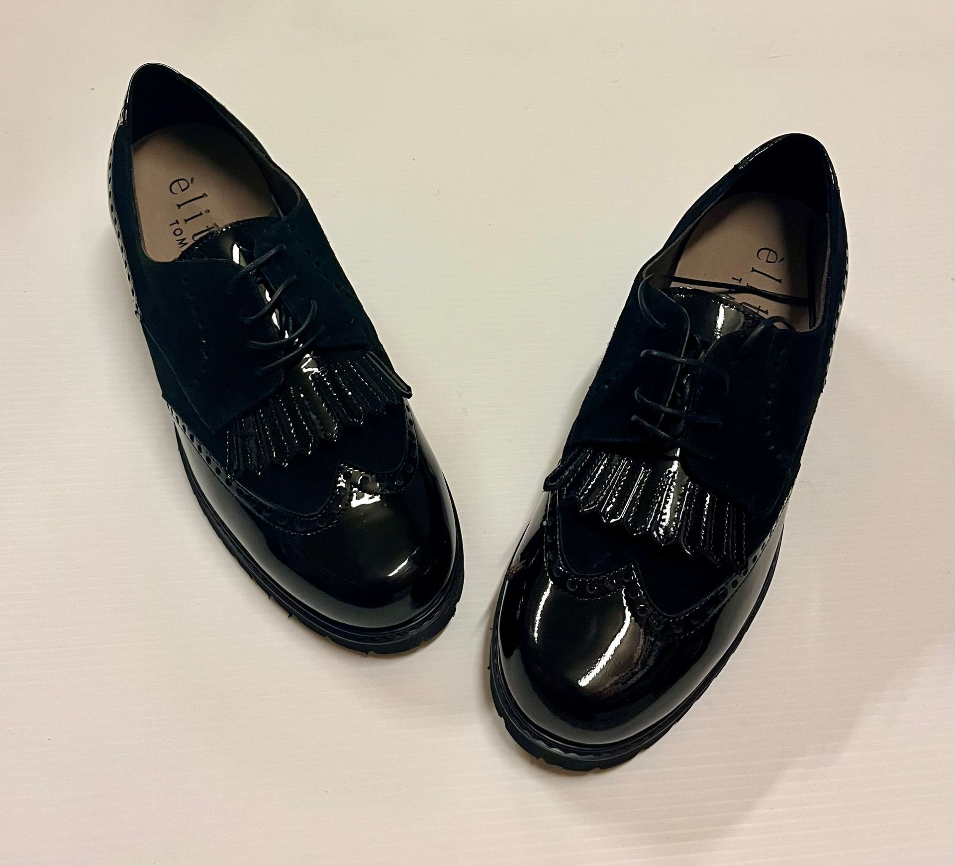 Scarpe stringate nere in vernice e pelle scamosciata con nappe su superficie bianca.