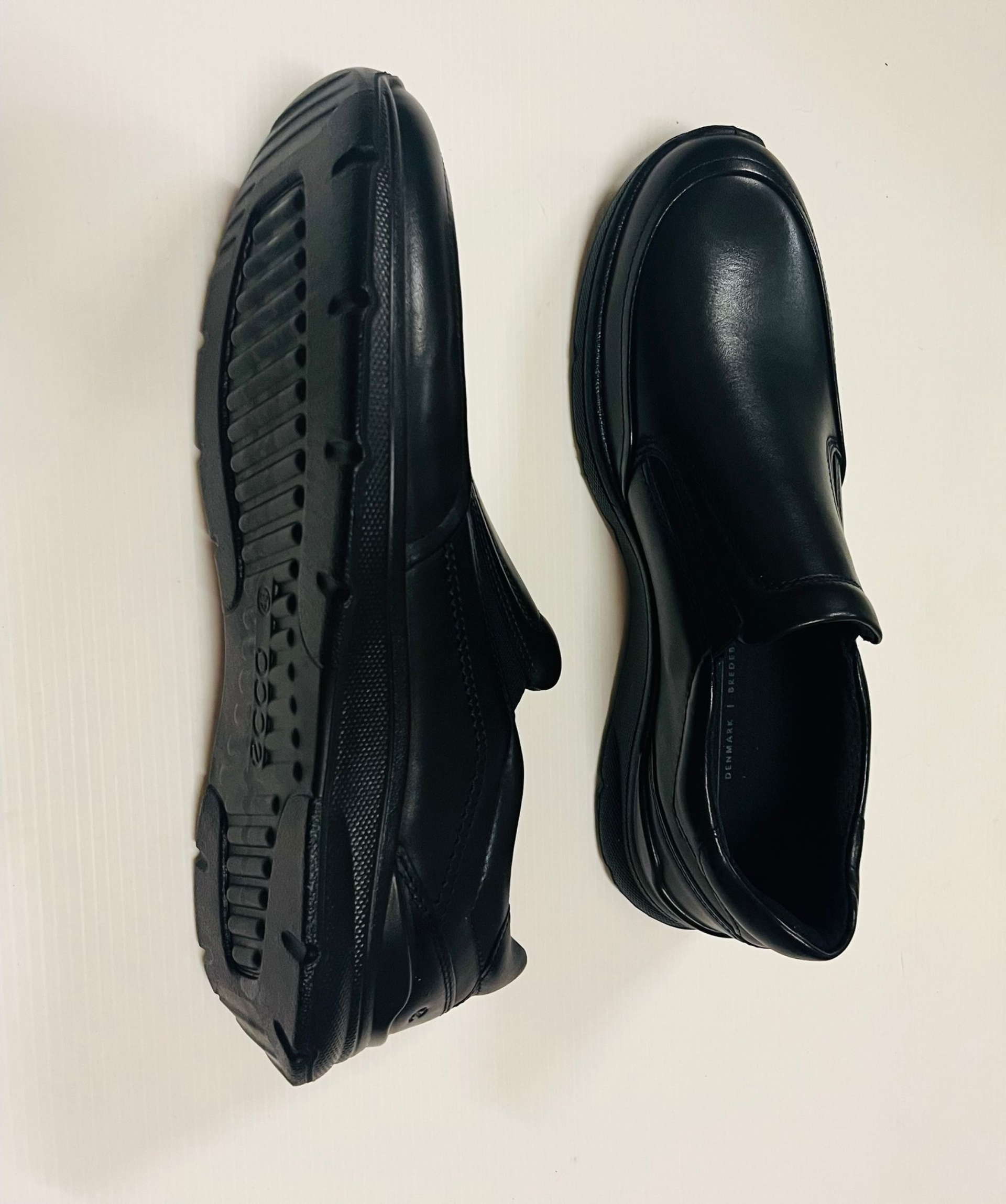 Due scarpe nere slip-on, una con la suola rivolta verso l'alto, su una superficie bianca.