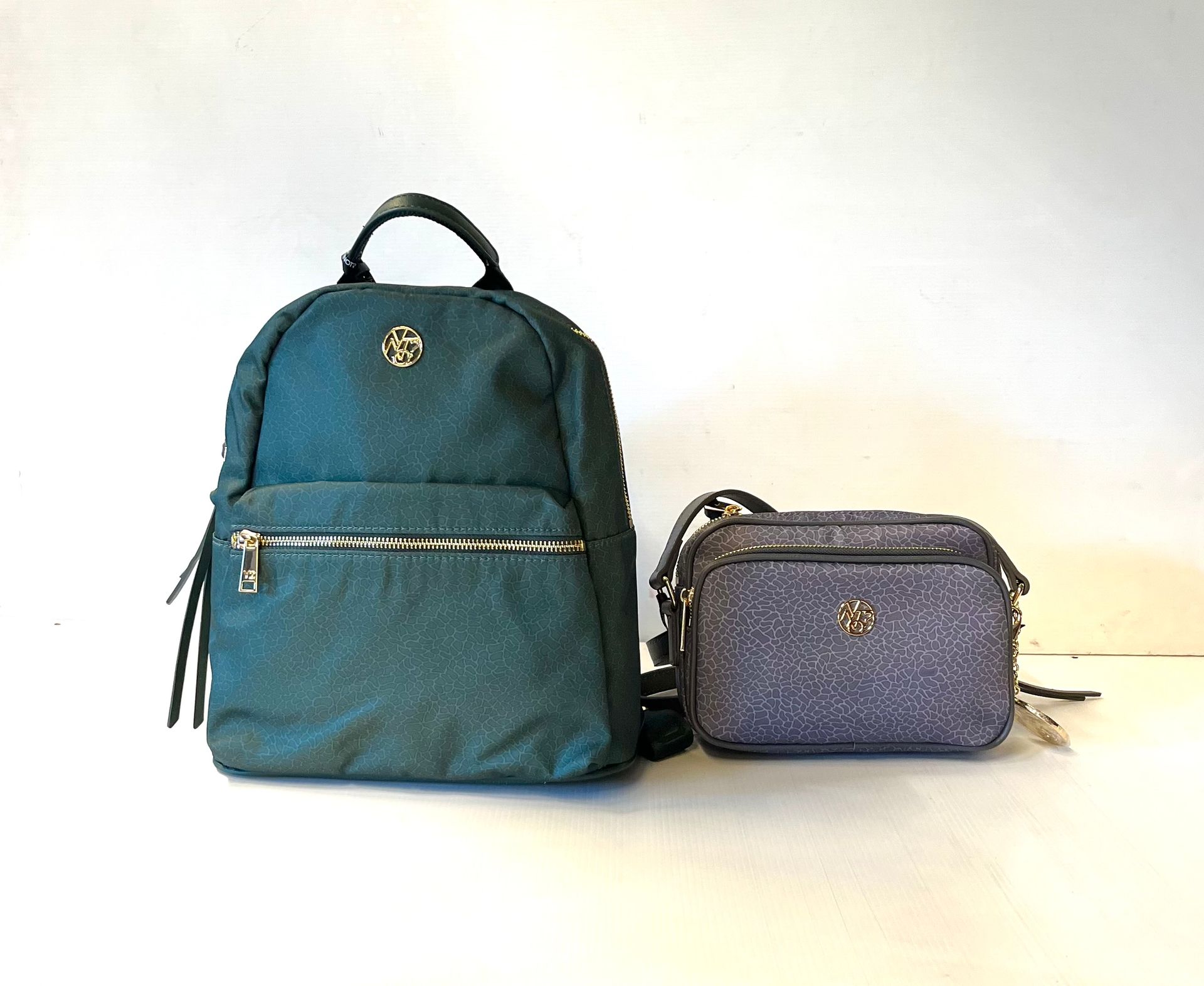 Zaino verde acqua e borsa a tracolla grigia glitterata con dettagli dorati su una superficie bianca.