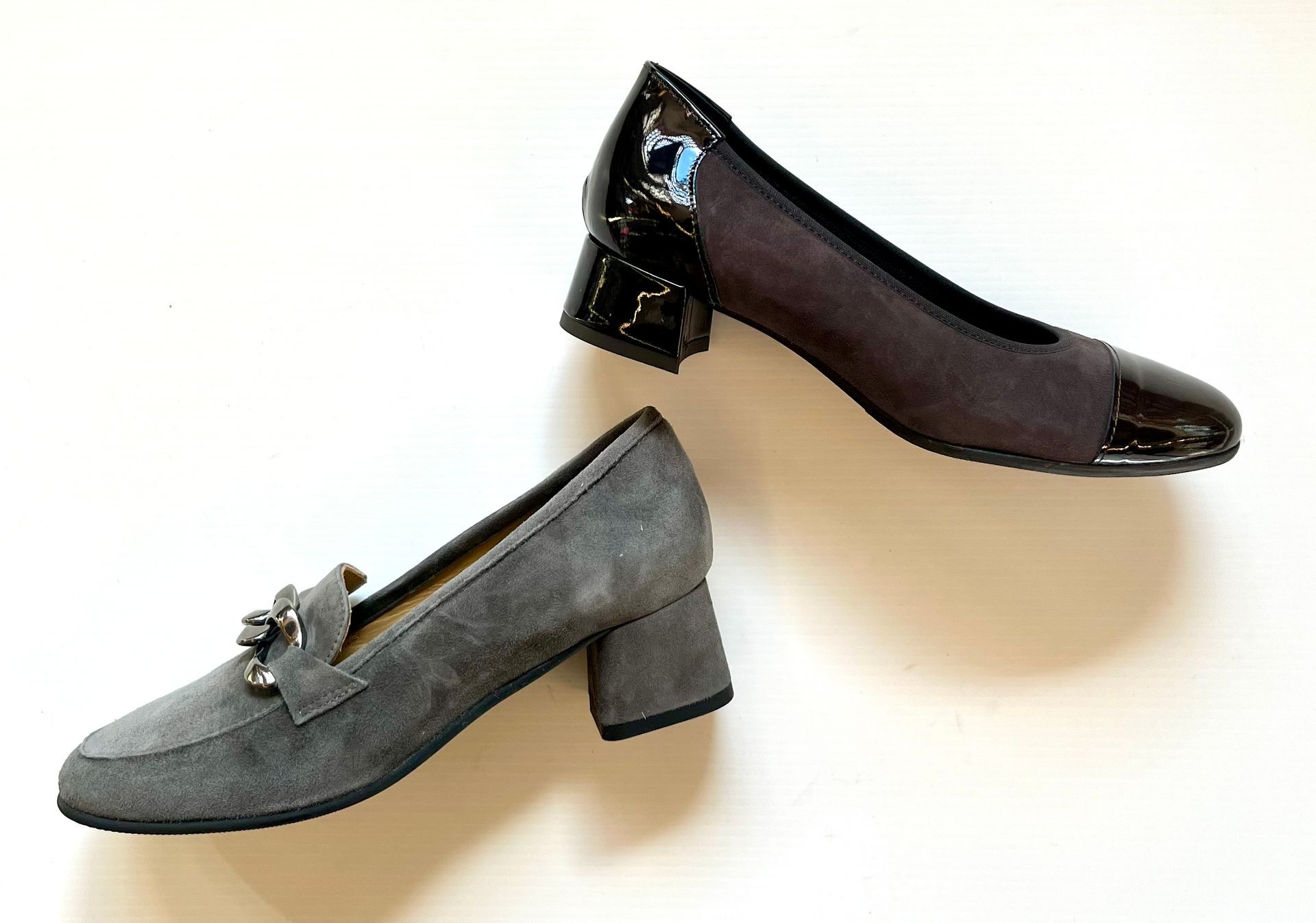 Due paia di scarpe eleganti da donna su superficie bianca: mocassini in pelle scamosciata grigia, décolleté nere e grigie, entrambe con tacco a blocco.