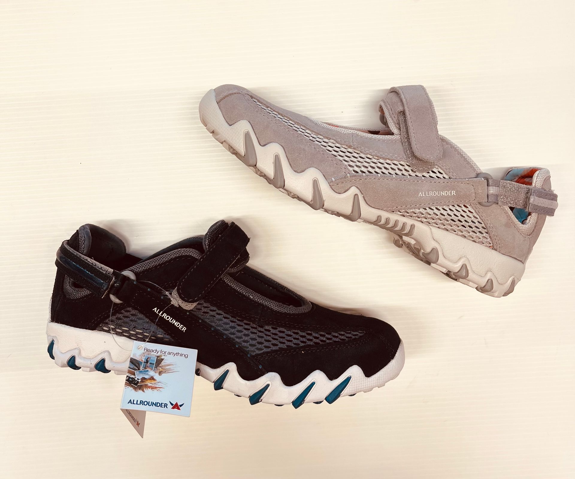 Due scarpe da acqua, una nera e una grigia, su sfondo crema. Entrambe hanno la suola ondulata e il cinturino in velcro.