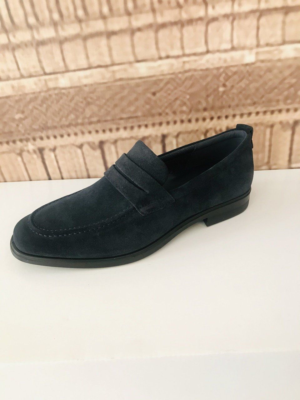 Mocassino in pelle scamosciata blu su una superficie bianca, con sfondo di un muro di mattoni.
